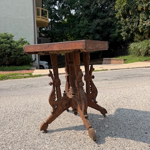 May include: A square wooden side table with a flat top and a central pedestal base. The base features four ornate, carved legs with decorative scrollwork and small wheels attached to the bottom of each foot, finished in a medium brown wood tone.