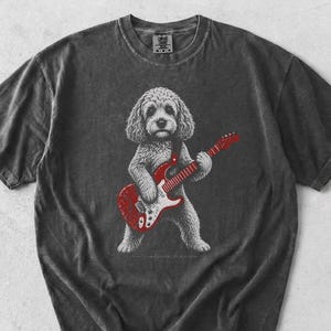 May include: A charcoal grey short-sleeved t-shirt featuring a graphic print of a fluffy dog standing on its hind legs and playing a bright red electric guitar. The shirt has a crew neckline and a vintage-style washed fabric texture.