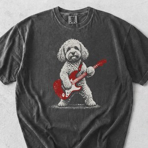 May include: A charcoal gray short-sleeve t-shirt featuring a white graphic illustration of a curly-haired dog standing on its hind legs while playing a red electric guitar. The shirt has a crew neckline and a vintage-style washed fabric finish.