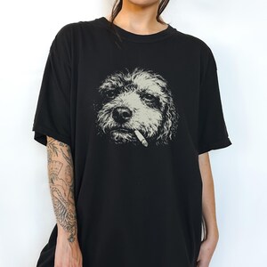 May include: A black short-sleeved t-shirt featuring a graphic print of a dog with curly fur. The dog is depicted in a monochromatic light grey tone and appears to have a cigarette held in its mouth. The design is centred on the chest area.