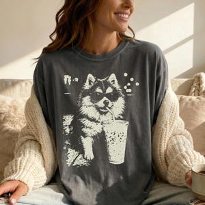 Op de afbeelding: Donkergrijs t-shirt met een zwart-witte illustratie van een hond met een zonnebril die uit een beker met een rietje drinkt. De hondenafbeelding staat in het midden van het shirt. Het shirt heeft een relaxte pasvorm.