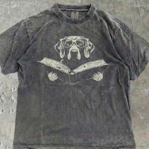 May include: A charcoal grey short-sleeve t-shirt with a mineral wash finish. The centre graphic features a line art illustration of a dog wearing glasses while reading an open book, with two paws visible at the bottom edges of the book pages.