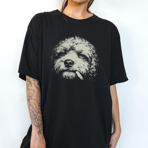 May include: A black short-sleeved t-shirt featuring a central graphic print of a curly-haired dog with a cigarette in its mouth. The dog illustration is rendered in shades of light grey and white, contrasting against the solid black fabric.