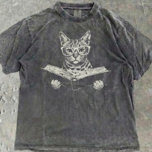 May include: A charcoal gray short-sleeve t-shirt with a mineral wash finish. The center of the shirt features a white graphic illustration of a cat wearing thick-rimmed glasses while holding an open book with both front paws.