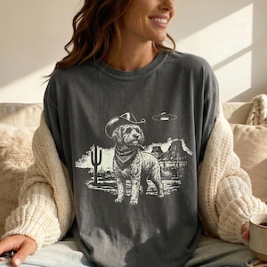 Peut inclure: T-shirt gris foncé avec un motif d'un chien portant un chapeau de cowboy et un bandana, sur un paysage désertique avec un cactus et des montagnes. Un OVNI plane au-dessus du chien. Le t-shirt est de coupe ample.