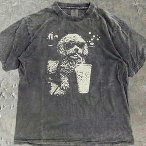 Könnte beinhalten: Dunkelgraues T-Shirt mit einem weißen Aufdruck eines Hundes mit Sonnenbrille, der aus einem Becher mit Strohhalm trinkt. Das Shirt hat einen Rundhalsausschnitt und kurze Ärmel. Der Stoff hat einen verblichenen Vintage-Look.