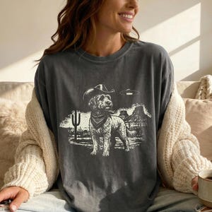 Può includere: T-shirt grigio scuro con un cane con cappello da cowboy e bandana, ambientato in un paesaggio desertico con un cactus e montagne. Un UFO fluttua sopra il cane. La maglietta ha una vestibilità ampia.
