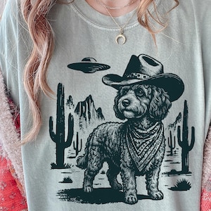 Peut inclure: Un t-shirt vert sauge avec une impression graphique noire représentant un chien aux poils bouclés portant un chapeau de cowboy et un bandana à motifs. Le motif montre un paysage désertique avec des cactus, des montagnes et une soucoupe volante.
