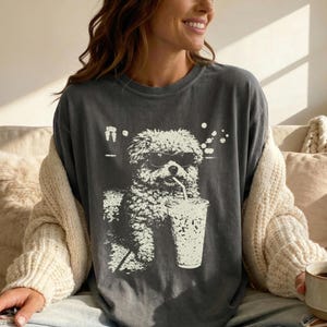 Pode incluir: Camiseta cinza escuro com um gráfico branco de um cachorro usando óculos de sol e bebendo de um copo com canudo. O cachorro é uma mistura de poodle. A camisa tem um ajuste folgado.