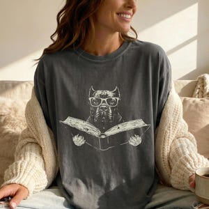 May include: A charcoal grey crew neck t-shirt featuring a white graphic print of a dog wearing glasses and reading an open book. The shirt has a relaxed fit and a vintage-style washed appearance, with the dog illustration centred on the chest.