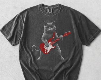 May include: A charcoal gray short-sleeve t-shirt featuring a graphic print of a cat playing a red electric guitar. The shirt has a crew neckline and a vintage-style washed finish. The cat is depicted in a stippled, monochromatic style.