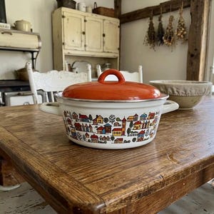 Könnte beinhalten: Ein Vintage-Emaille-Topf mit orangefarbenem Deckel und weißem Körper. Der Topf ist mit einer farbenfrohen Dorfszene verziert. Er hat zwei Seitengriffe und steht auf einem Holztisch. Eine charmante Ergänzung für jede Küche.
