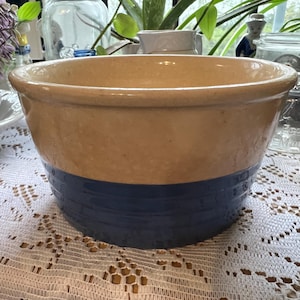May include: A vintage stoneware mixing bowl with a two-tone design. The upper portion is a light tan color, while the lower section features a deep blue band. The bowl has a wide, rounded shape and a thick rim, suggesting it's a durable kitchen item.