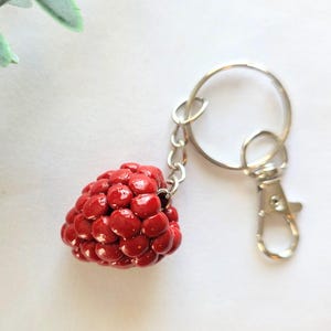 May include: A red raspberry-shaped keychain with a silver-colored chain and clasp. The keychain is made of a glossy, textured material, and the raspberry has small white accents. The background is a soft white with a hint of green foliage.