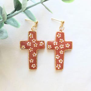 May include: A pair of cross-shaped earrings. The earrings are a terracotta color with a gold border and small white floral details. The earrings are hanging from gold posts.