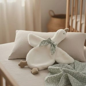 May include: A plush white goose toy with a green floral bow and feet, resting on a beige pillow and blanket. The nursery setting includes a crib and a woven basket.