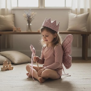 May include: A child's fairy costume set, including a pink crown, wings, and wand. The crown and wings are patterned with small white dots. The child is wearing a pink long-sleeved shirt and shorts.