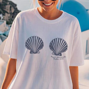 Puede incluir: Camiseta blanca con dos dise&ntilde;os de conchas marinas grises. El texto en la camiseta dice "Megan's Bachelorette, Follia, 2024". La camiseta es una impresi&oacute;n de mezclilla.