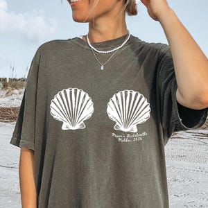 Puede incluir: Camiseta verde oliva oscuro con un dise&ntilde;o de concha blanca. La camiseta tiene el texto "Pepper's Bachelorette, Malibu, 2026". La camiseta es de corte holgado y se lleva en la playa.