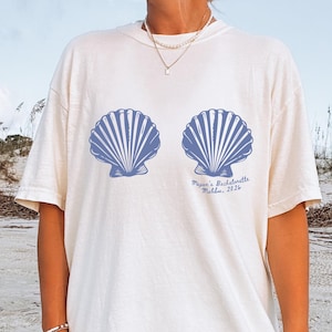 Puede incluir: Camiseta color marfil con dos diseños de conchas marinas azules. La camiseta incluye el texto "Megan's Bachelorette Malibu, 2026" en una fuente cursiva. Es una prenda informal con temática playera.