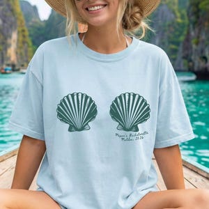 Puede incluir: Camiseta azul claro de chambray con dise&ntilde;o de concha azul abeto. La camiseta tiene el texto "Megan's Bachelorette Malibu, 2326" impreso. Corte holgado.