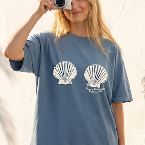 May include: Blue jean t-shirt with ivory seashell print. The shirt features two large seashell graphics and the text "Megan's Bachelorette Malibu, 2026". The shirt is a casual, comfortable style.