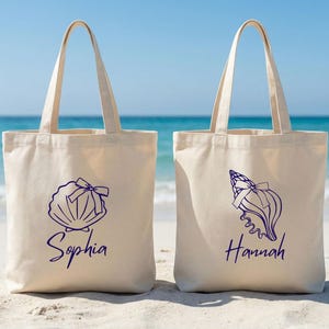 以下が含まれることがあります： 紺色の貝殻のデザインと名前が入った、ベージュ色のキャンバス地のトートバッグが2つ。片方のバッグには「Sophia」、もう片方には「Hannah」と書かれています。バッグには長い持ち手があり、砂浜に置かれ、背景には海が広がっています。