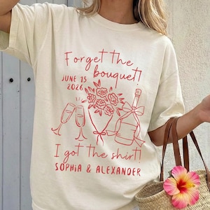 Op de afbeelding: Ivoor t-shirt met karmozijnrode print. Het ontwerp bevat de tekst "Forget the bouquet!" en "I got the shirt!" samen met een datum, champagneglazen, een boeket en een champagnefles. De namen Sophia & Alexander staan ook op het shirt.
