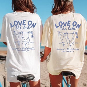 May include: Two short-sleeved t-shirts, one white and one ivory, featuring a blue graphic on the back. The design includes the text 'LOVE ON the lake', 'est. 2026', 'Lauren's Bachelorette', and 'LAKE TAHOE, CA' with a clothesline illustration.