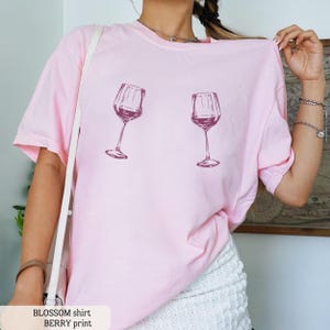 May include: A light pink t-shirt featuring two wine glasses filled with a dark red liquid. The shirt has short sleeves and a relaxed fit. The text "BLOSSOM shirt BERRY print" is at the bottom of the image.