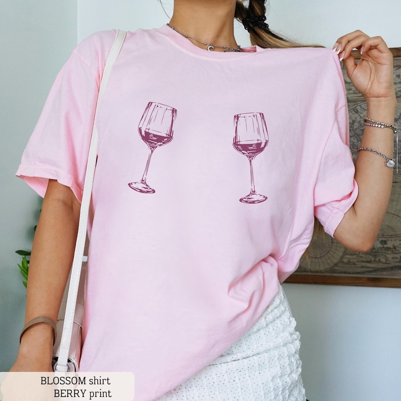 May include: A light pink t-shirt featuring two wine glasses filled with a dark red liquid. The shirt has short sleeves and a relaxed fit. The text "BLOSSOM shirt BERRY print" is at the bottom of the image.