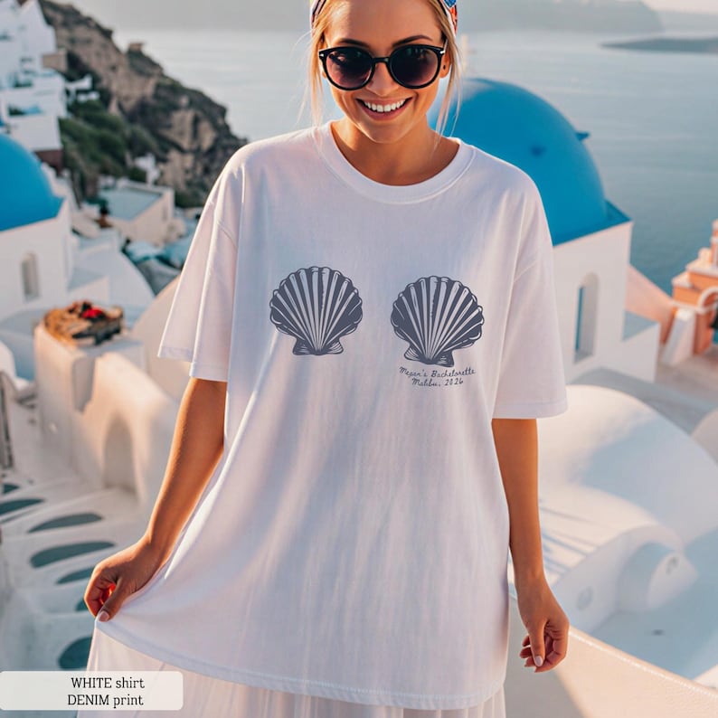 Puede incluir: Camiseta blanca con dos dise&ntilde;os de conchas marinas grises. El texto en la camiseta dice "Megan's Bachelorette, Follia, 2024". La camiseta es una impresi&oacute;n de mezclilla.