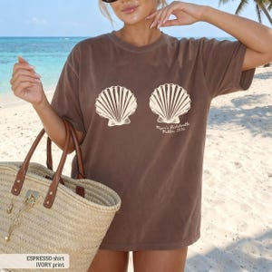Puede incluir: Camiseta color espresso con gr&aacute;fico de concha de color marfil. La camiseta presenta dos dise&ntilde;os grandes de conchas marinas y el texto "Megan's Bachelorette Malibu, 2026". La camiseta se usa en una playa.
