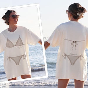 K&ouml;nnte beinhalten: Ein elfenbeinfarbenes T-Shirt mit einem espressofarbenen Grafikdruck. Die Vorderseite zeigt ein Bikini-Oberteil und -Unterteil mit dem Text "Ko We Fi". Die R&uuml;ckseite zeigt die Ansicht eines Bikinis mit dem Text "Bridesmaid" unter der Schleife.