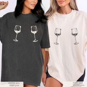 May include: Two t-shirts, one pepper-colored and one ivory, each featuring a wine glass graphic. The pepper shirt has an ivory print, and the ivory shirt has a pepper print. The shirts are short-sleeved and have a relaxed fit.