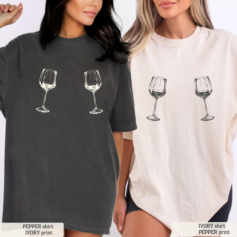 May include: Two t-shirts, one pepper-colored and one ivory, each featuring a wine glass graphic. The pepper shirt has an ivory print, and the ivory shirt has a pepper print. The shirts are short-sleeved and have a relaxed fit.