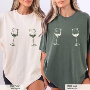 May include: Two t-shirts, one ivory and one moss green, each featuring a sketch of two wine glasses. The ivory shirt has the text "IVORY shirt, MOSS print." The moss green shirt has the text "MOSS shirt, IVORY print."