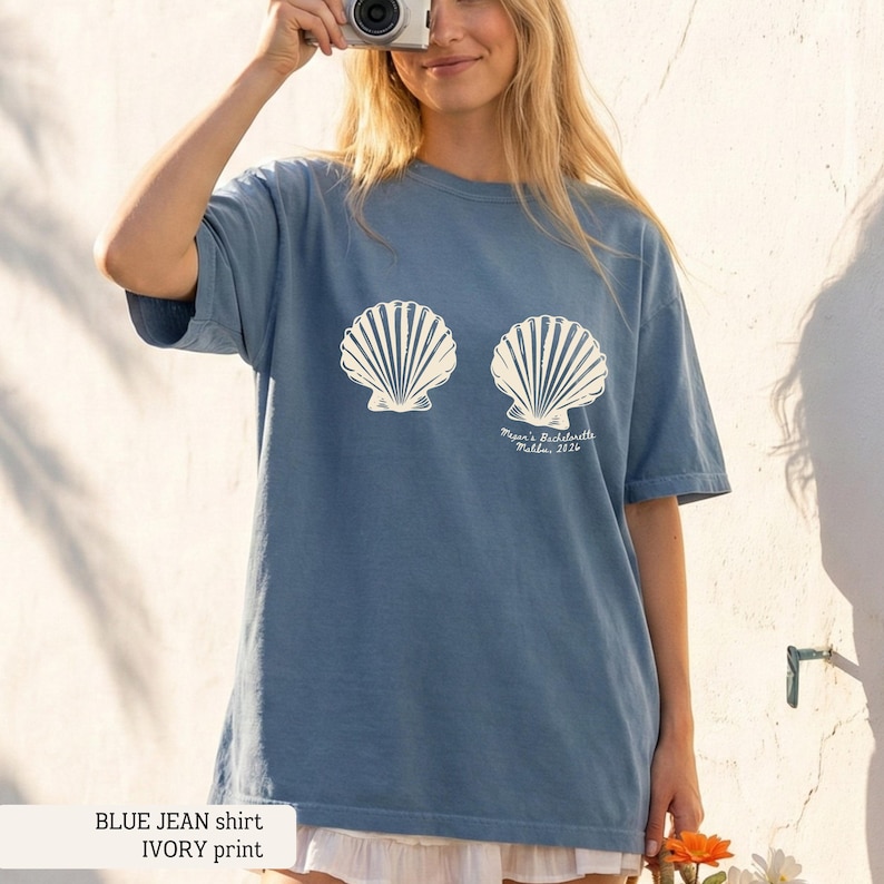 May include: Blue jean t-shirt with ivory seashell print. The shirt features two large seashell graphics and the text "Megan's Bachelorette Malibu, 2026". The shirt is a casual, comfortable style.