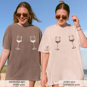 May include: Two women wearing oversized t-shirts with a wine glass graphic. One shirt is espresso-colored with an ivory print, and the other is ivory with an espresso print. The shirts have short sleeves and a relaxed fit.