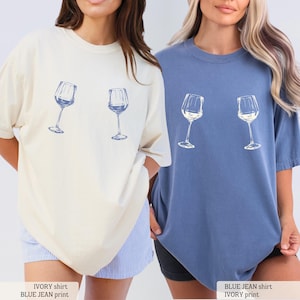 May include: Two oversized t-shirts, one ivory and one blue jean, each featuring a blue print of two wine glasses. The shirts have a relaxed fit and short sleeves. The text at the bottom reads "IVORY shirt BLUE JEAN print" and "BLUE JEAN shirt IVORY print".