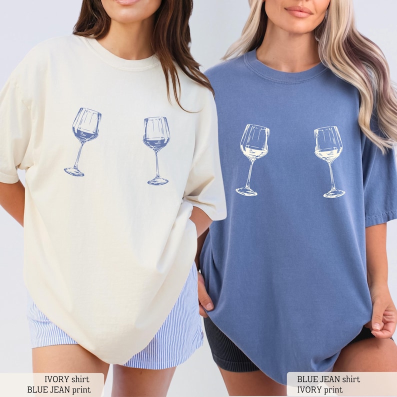 May include: Two oversized t-shirts, one ivory and one blue jean, each featuring a blue print of two wine glasses. The shirts have a relaxed fit and short sleeves. The text at the bottom reads "IVORY shirt BLUE JEAN print" and "BLUE JEAN shirt IVORY print".