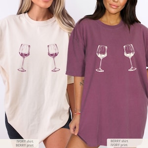 May include: Two oversized t-shirts, one ivory and one berry, each featuring a wine glass graphic. The ivory shirt has a berry print, and the berry shirt also has an ivory print. The shirts are casual and comfortable, perfect for everyday wear.