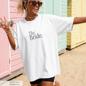 Puede incluir: Camiseta blanca con el texto "The Bride" en azul marino. La camiseta tiene un corte holgado y parece estar hecha de un material suave. La camiseta forma parte de una colección nupcial.