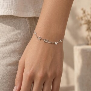 May include: A silver-toned chain bracelet featuring the name Olivia in cursive script, accented with a small five-petaled flower charm, a butterfly charm, and a delicate leaf-shaped charm integrated into the thin metal link chain design.