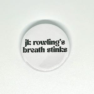 May include: A circular white pin-back button featuring black text that reads "jk rowling's breath stinks" in a bold, rounded, lowercase font. The button has a smooth, glossy finish and a simple, clean design with the text centred on the surface.