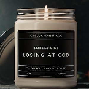 Può includere: Una candela profumata in un barattolo di vetro con coperchio argentato. L'etichetta recita "CHILLCHARM CO. SMELLS LIKE LOSING AT COD. IT'S THE MATCHMAKING'S FAULT." La candela è da 255 grammi e dura 60 ore.