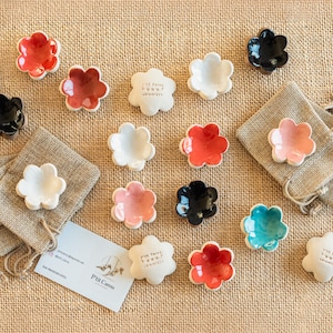May include: A selection of small, flower-shaped ceramic dishes in various colours, including red, black, white, pink, and teal. Some dishes are paired with small hessian bags. The dishes have a smooth, glossy finish and are arranged on a textured, tan surface.