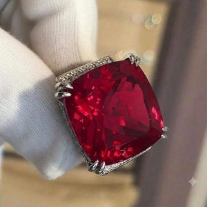 May include: A large cushion-cut deep red gemstone ring set in a silver-toned metal band. The setting features a decorative border of small clear stones along the edges and four prominent prongs securing the central red stone in place.
