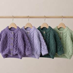 Könnte beinhalten: Vier langärmelige Strickjacken mit Zopfmuster, die an Holzkleiderbügeln hängen. Die Strickjacken sind in den Farben Dunkellila, Helllavendel, Dunkelwaldgrün und Blasssalbeigrün gehalten. Jede hat einen Rundhalsausschnitt, gerippte Bündchen und braune Knöpfe.