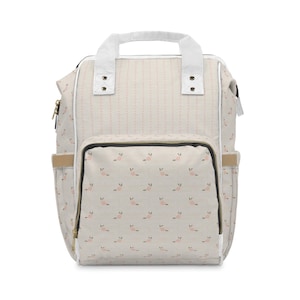 May include: A beige and white floral print backpack with a top handle and adjustable straps. The backpack features a front pocket with a floral pattern and a vertical dotted pattern on the main body. Gold zippers and white trim complete the design.